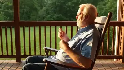 A man in his sixties in a rocking chair on a porch taking a small white spray bottle from his shirt pocket