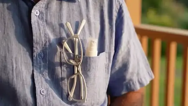 A man's hand drawing a small white spray bottle from his shirt pocket beside his reading glasses and shaking it